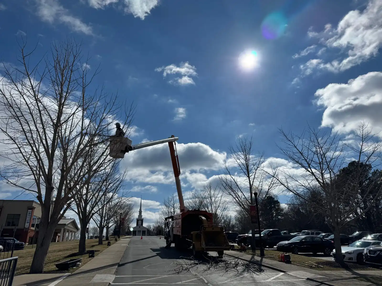 Greenbrier Tree Experts bucket truck with its boom raised into a bare street tree, a church steeple in the background